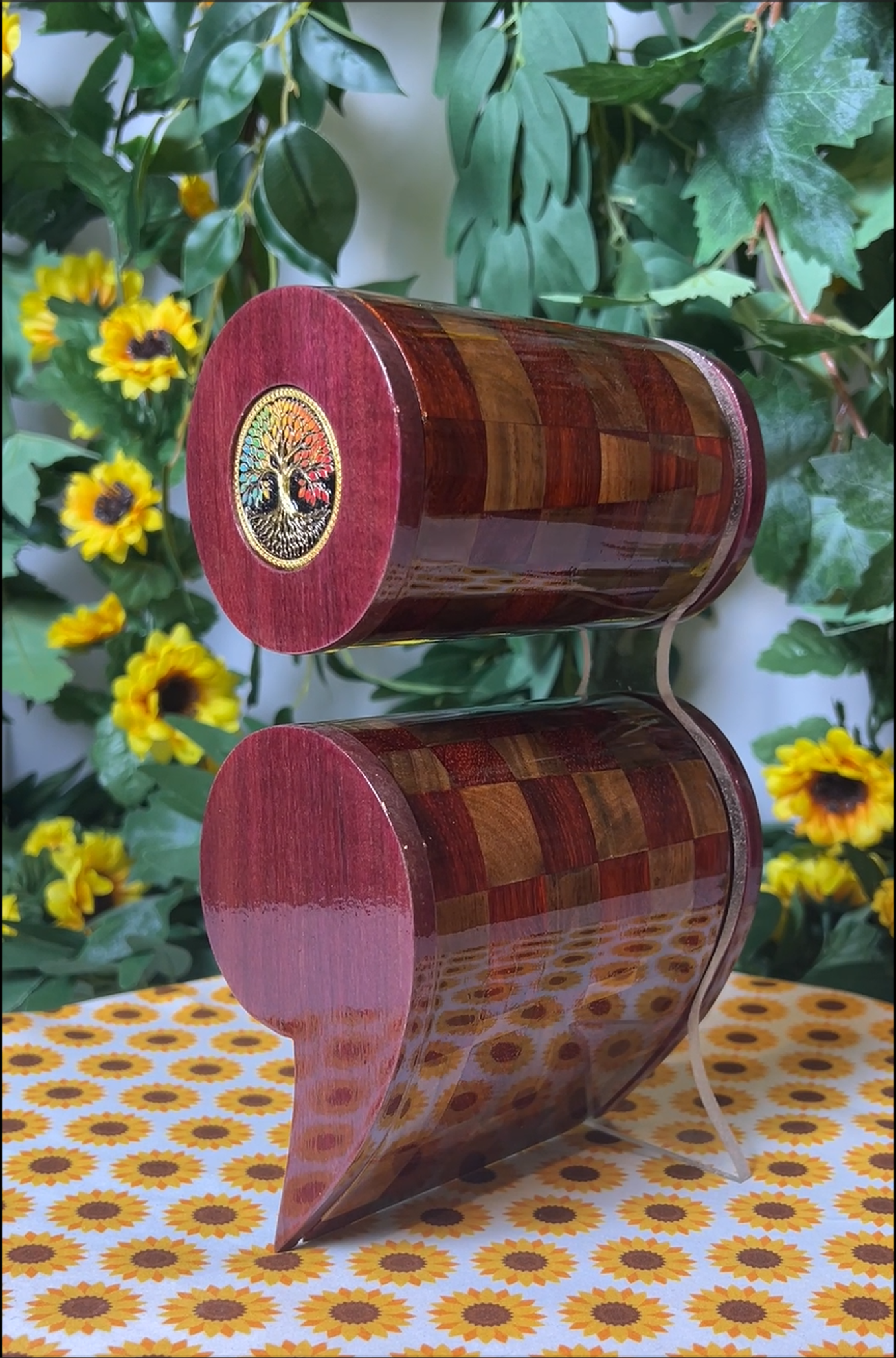 A wooden semicolon shaped box with a checkered pattern on a floral tablecloth with sunflowers and greenery in the background.