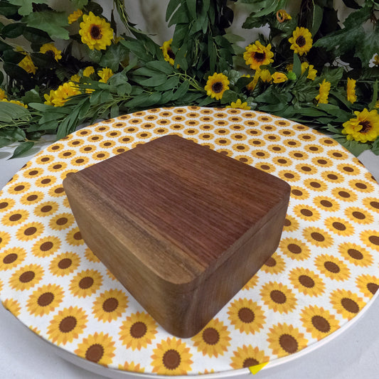Wooden box on a round table with sunflower pattern, surrounded by greenery and yellow flowers.