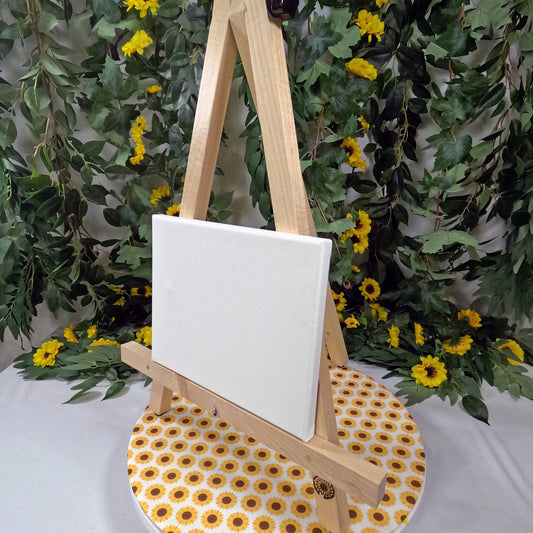 Wooden easel with a white canvas against a backdrop of greenery and yellow flowers.