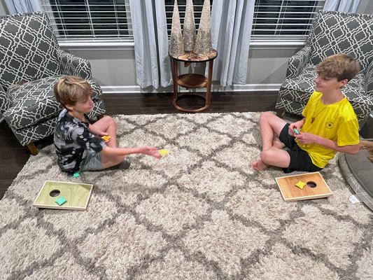 Two children playing cornhole on a carpeted floor with bean bags.