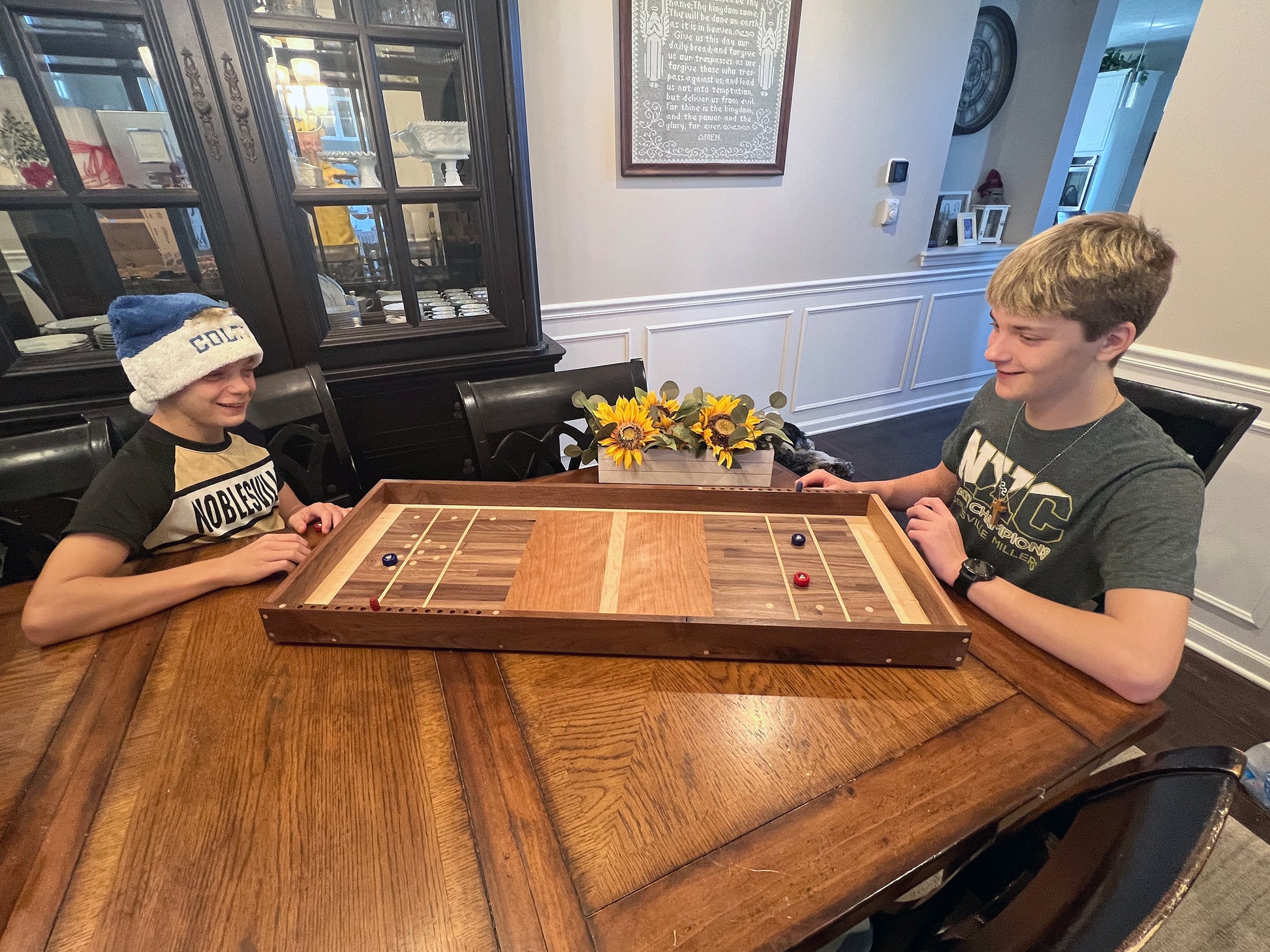 Two children playing a board game at a wooden table in a room with a bookshelf and decorative items.