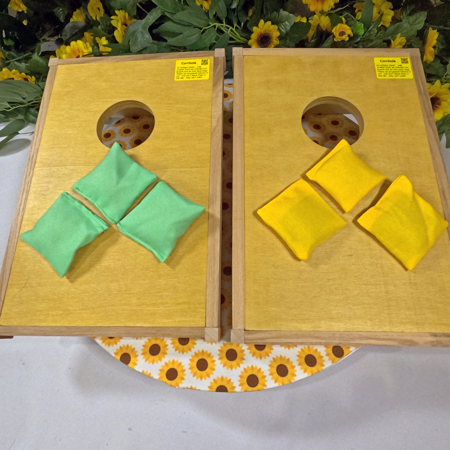 Two wooden cornhole boards with green and yellow bags on a white surface with sunflower pattern.