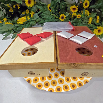 Wooden cornhole set with red and white boards on a white surface with sunflower pattern, surrounded by sunflowers.
