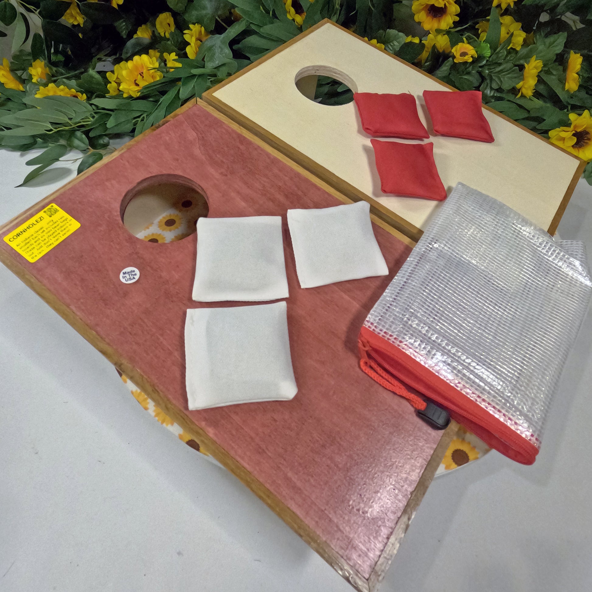 Two cornhole boards with bags on a white surface with greenery in the background