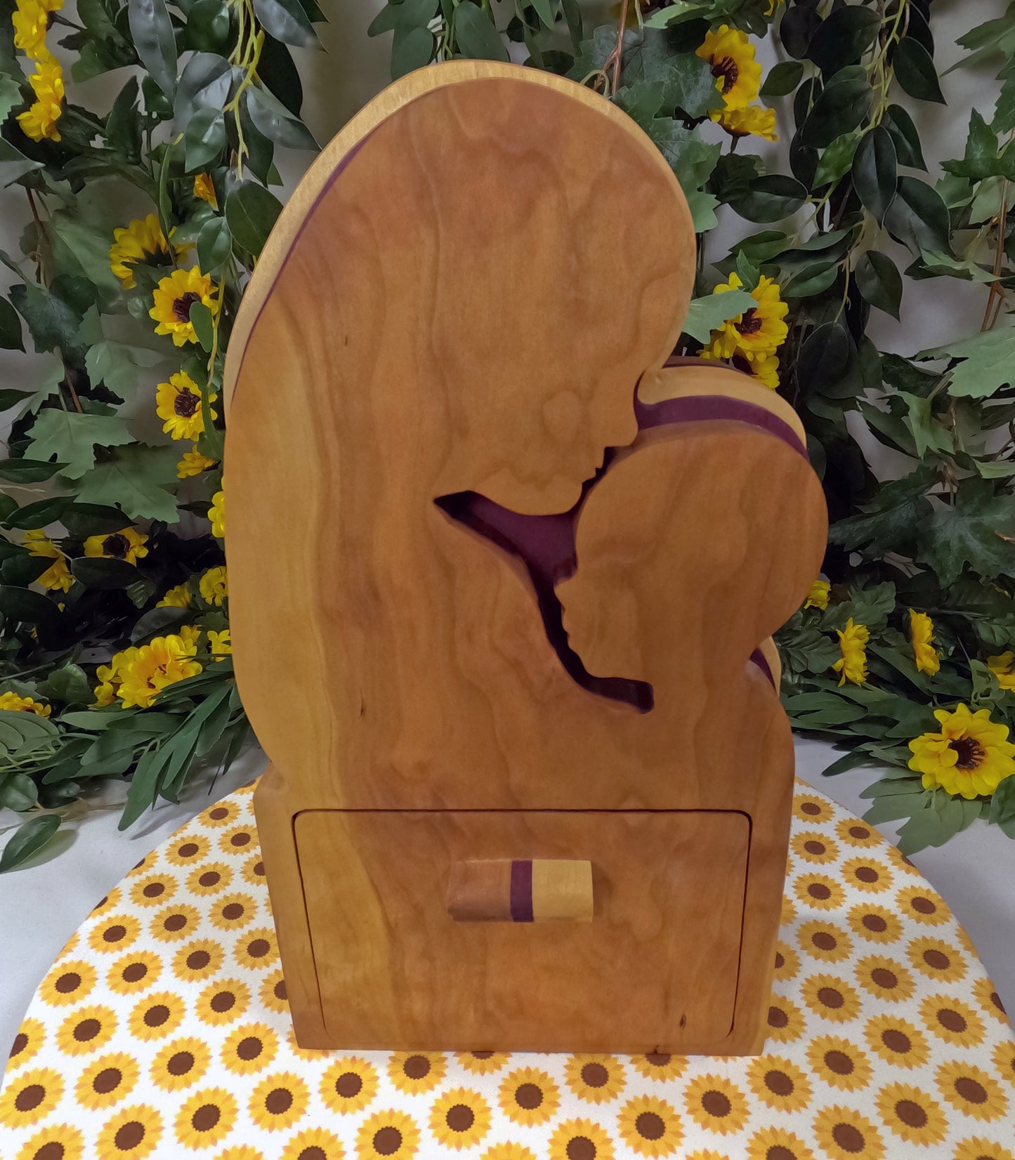 Wooden box with a cutout design of a person in a purple garment, placed on a floral-patterned surface with greenery and flowers in the background.