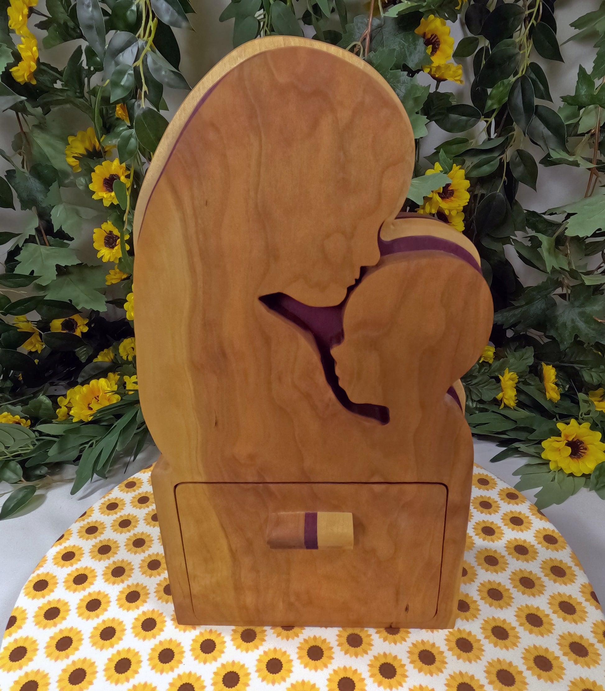 Wooden box with a cutout design of a person in a purple garment, placed on a floral-patterned surface with greenery and flowers in the background.