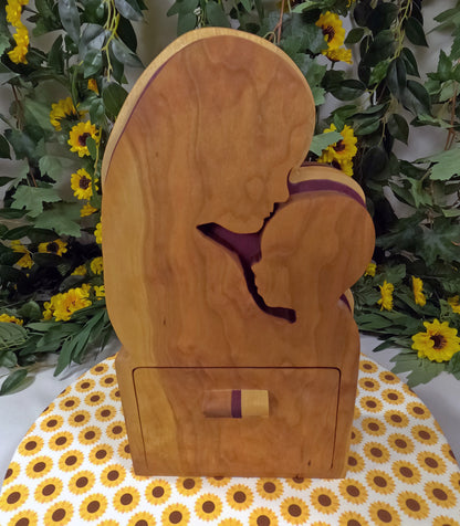 Wooden box with a cutout design of a person in a purple garment, placed on a floral-patterned surface with greenery and flowers in the background.