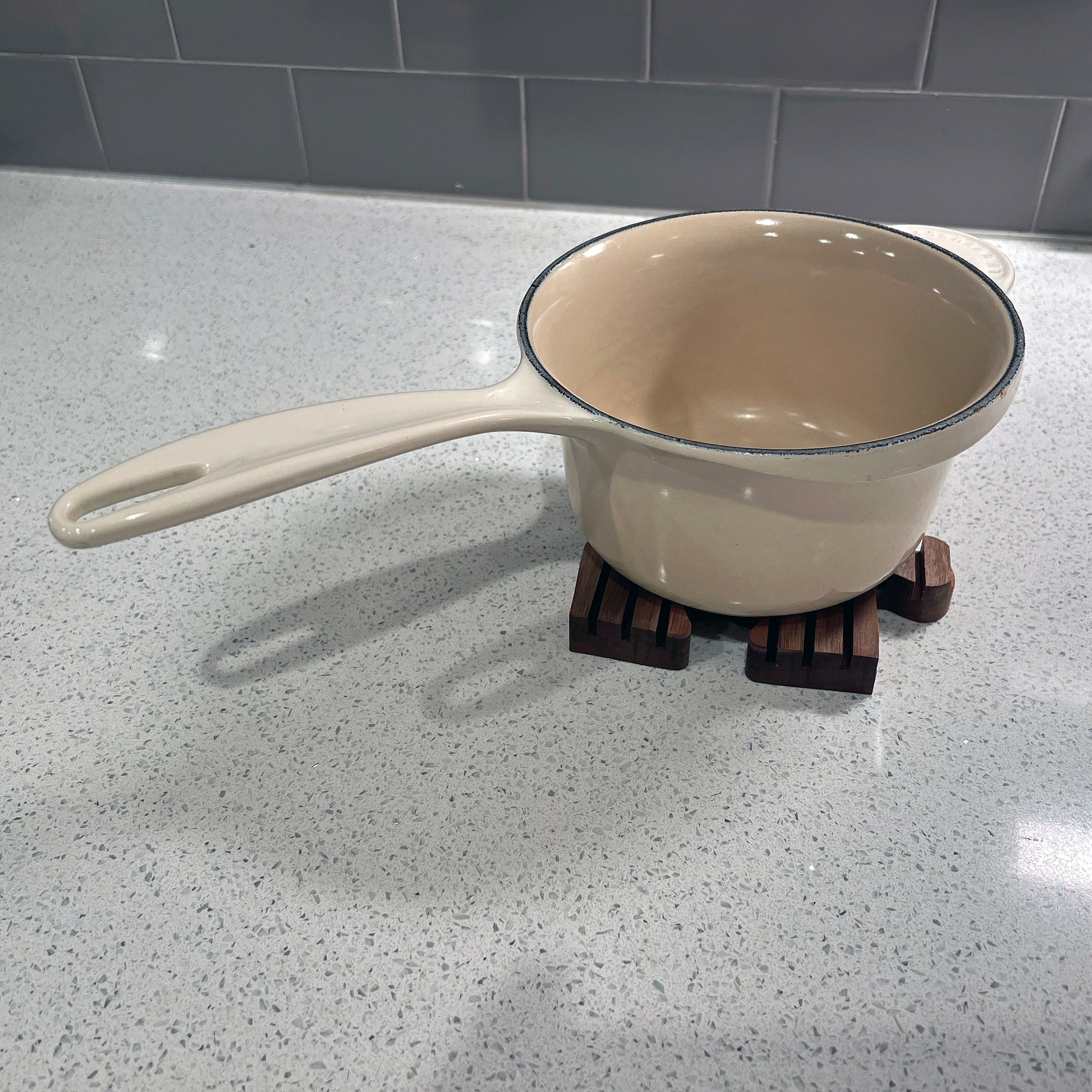 Beige saucepan on a wooden trivet on a speckled countertop with a tiled backsplash.