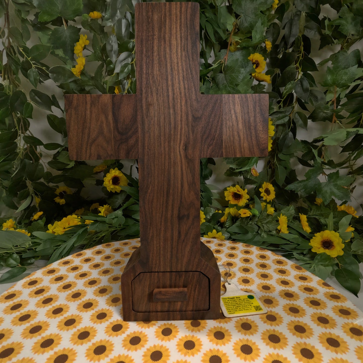 Wooden cross on a tablecloth with sunflower pattern, surrounded by greenery and flowers.