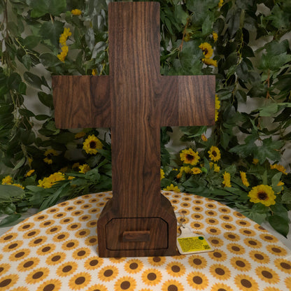 Wooden cross on a tablecloth with sunflower pattern, surrounded by greenery and flowers.
