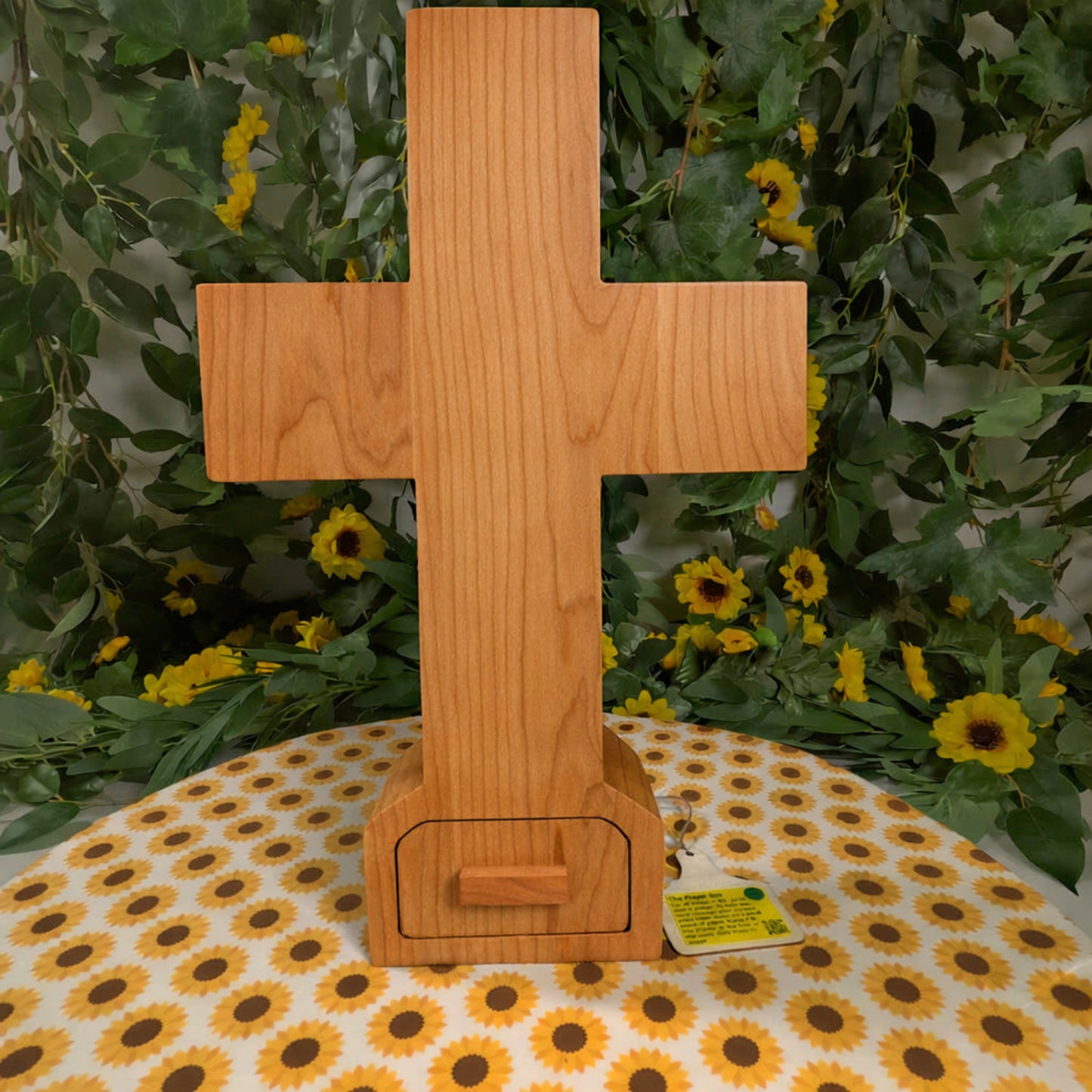 Wooden cross-shaped object on a tablecloth with sunflower pattern, against a green leafy background.