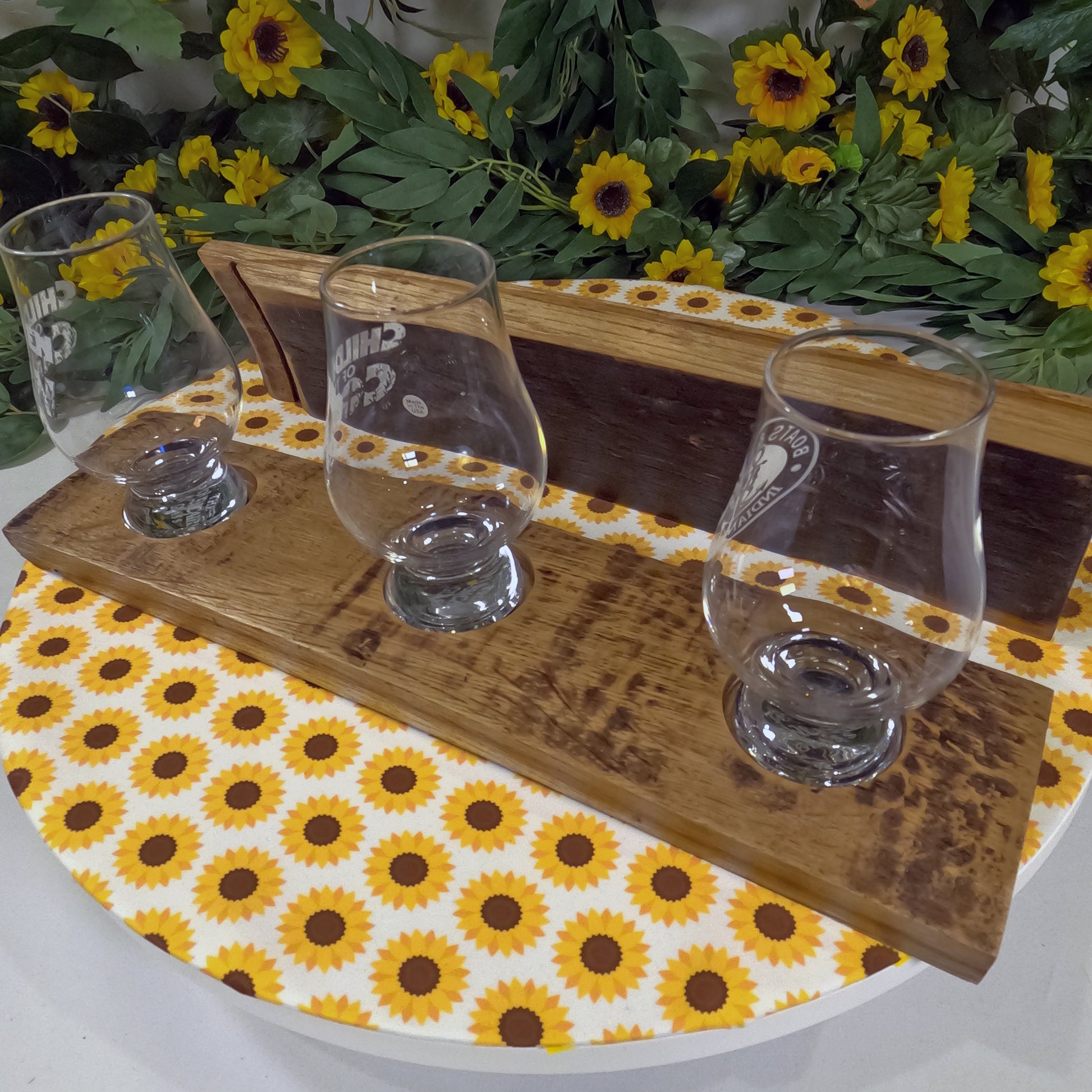 Three whiskey glasses on a wooden tray with sunflower design, surrounded by sunflowers and greenery.