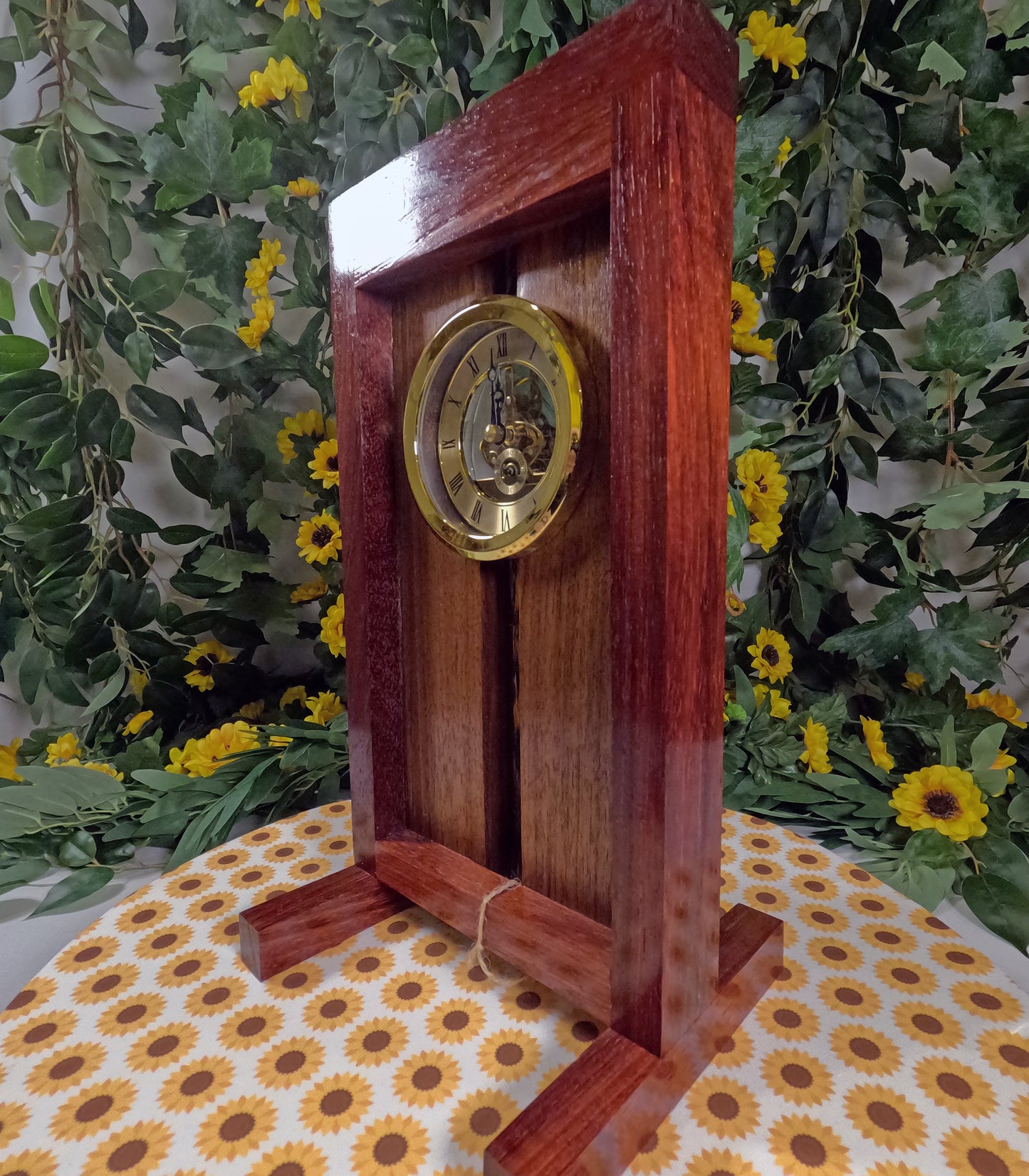 Wooden clock with gold face on a floral-patterned surface with greenery in the background