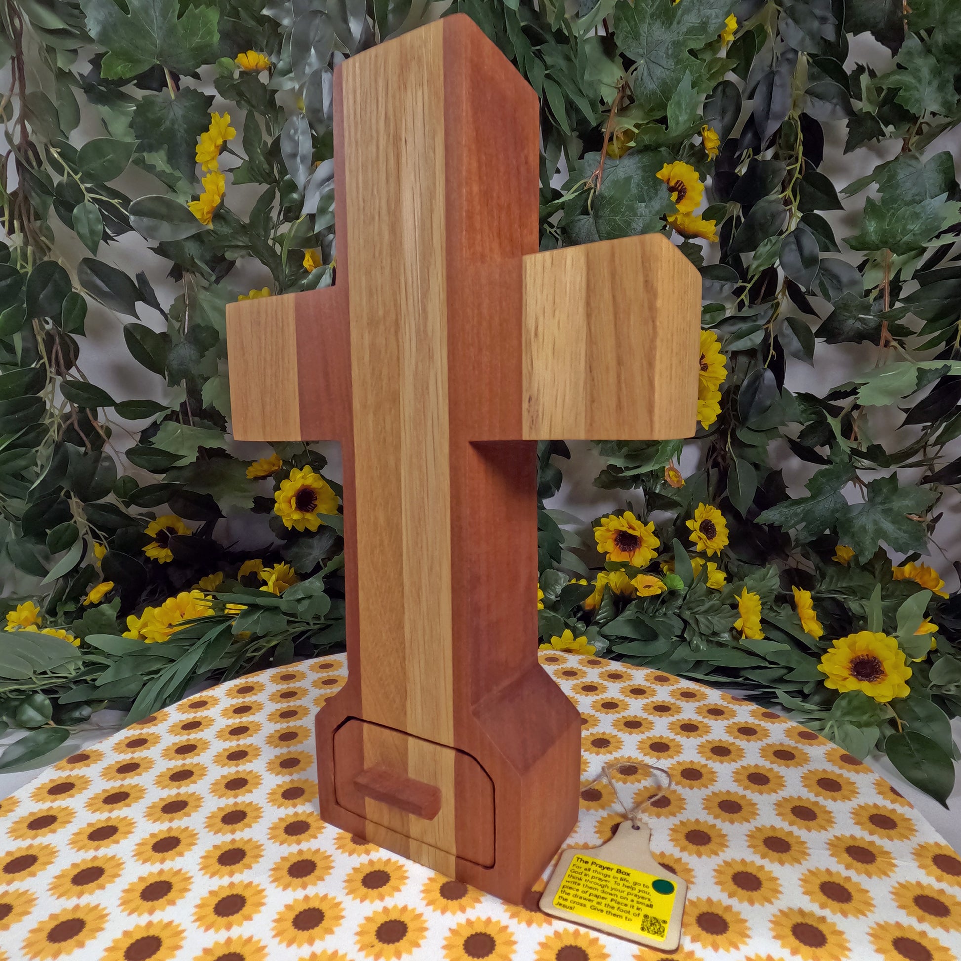 Wooden cross on a tablecloth with sunflower pattern, surrounded by greenery and flowers.