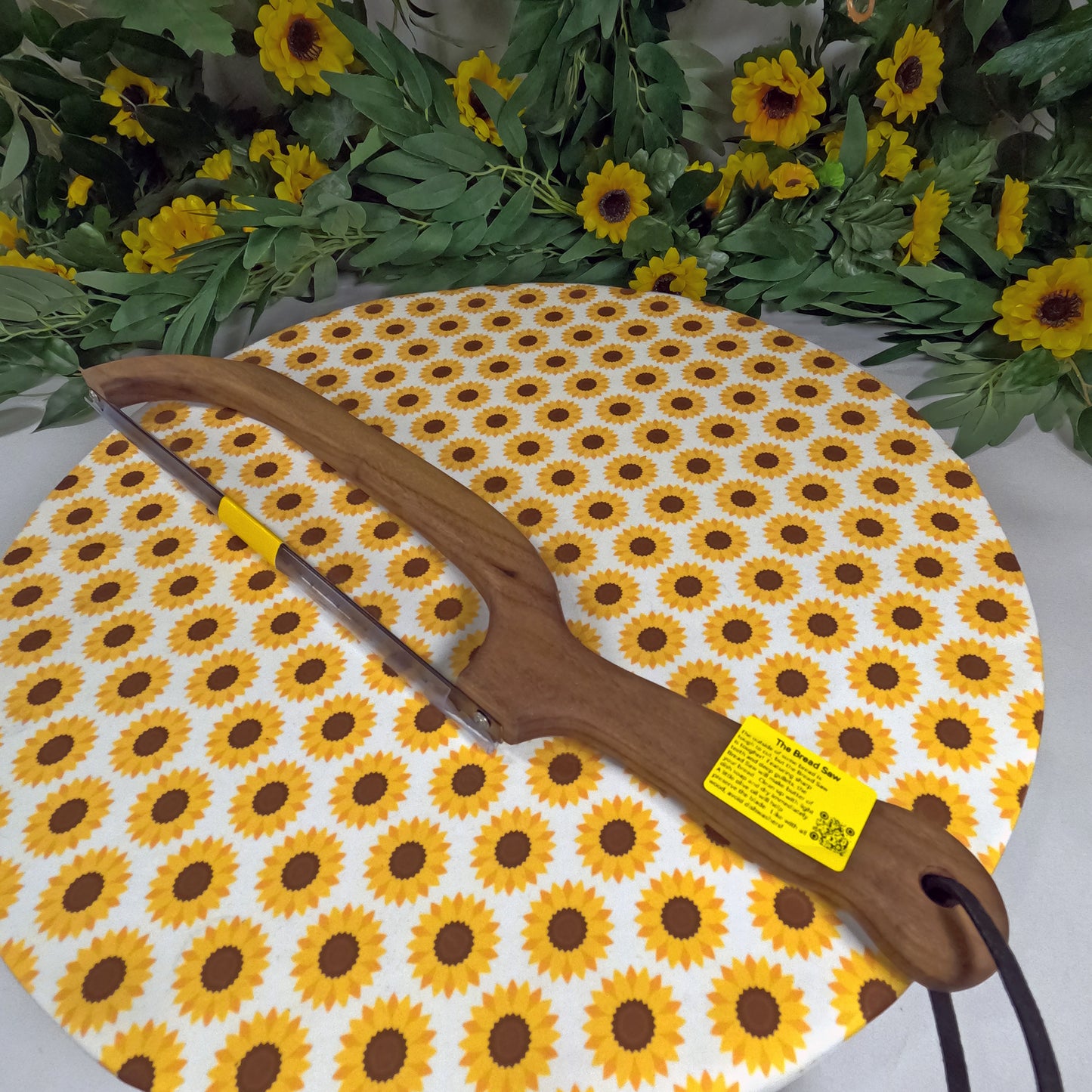 Wooden saw with a yellow label on a sunflower-patterned mat, surrounded by sunflowers and greenery.
