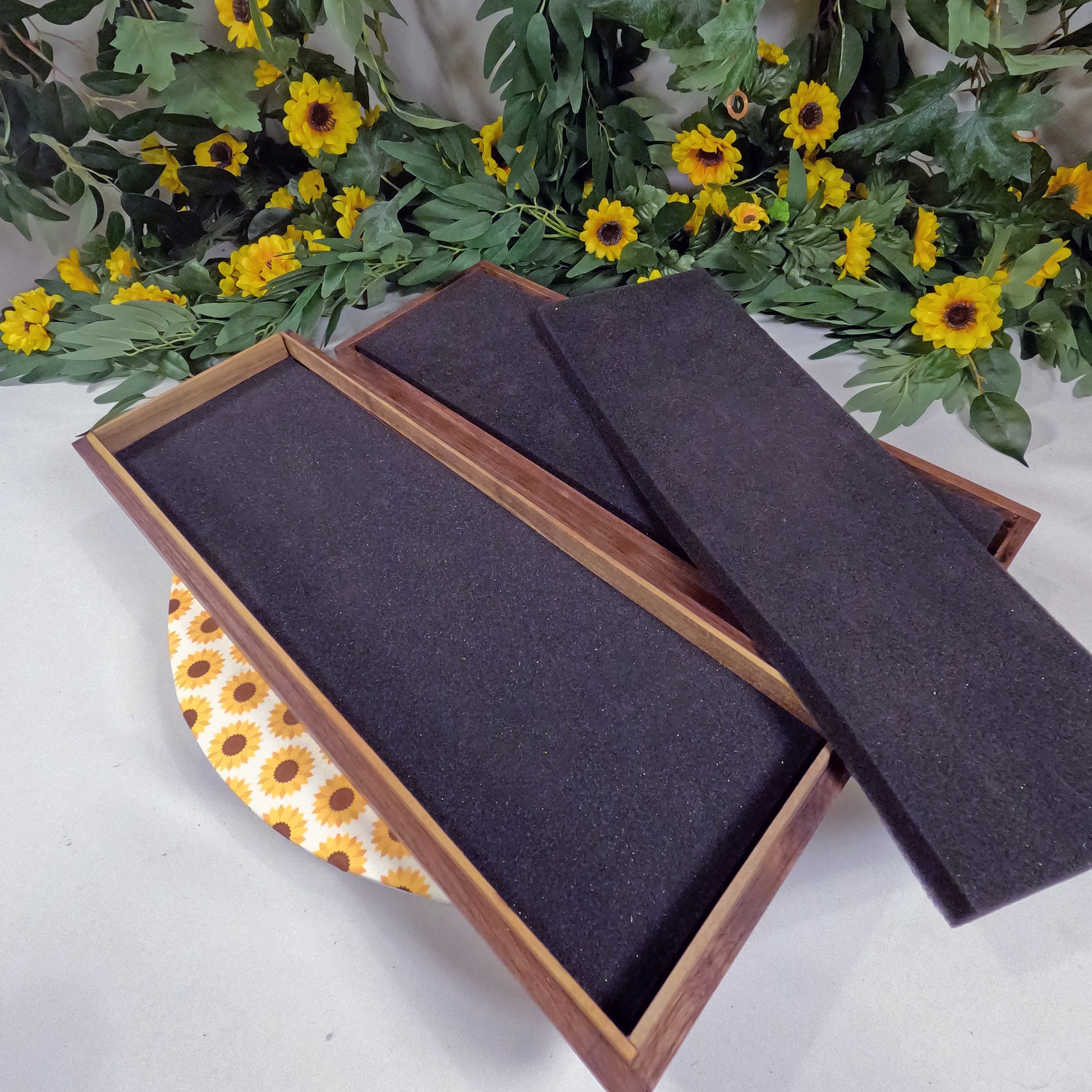 Wooden box with dark interior on a white surface with yellow flowers and green leaves in the background