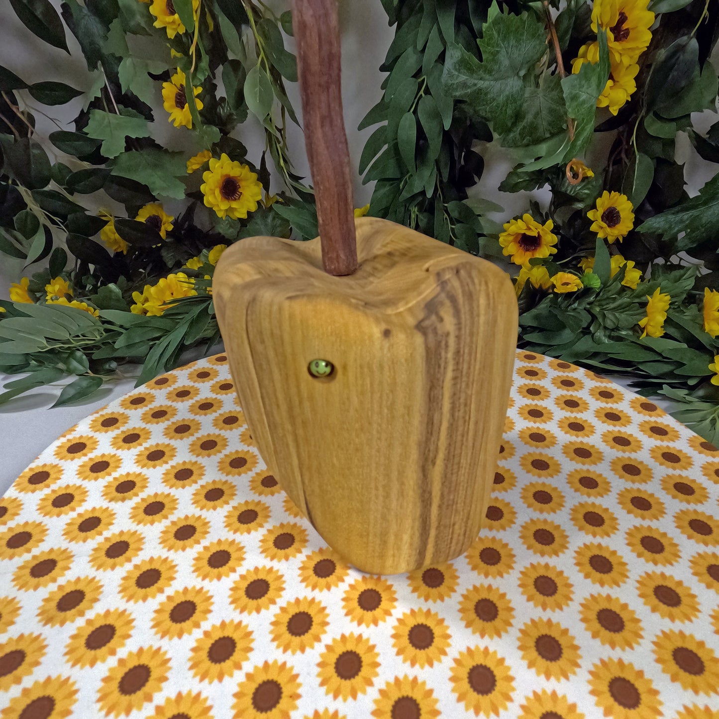 Wooden apple-shaped object on a tablecloth with sunflower pattern, surrounded by greenery and yellow flowers.