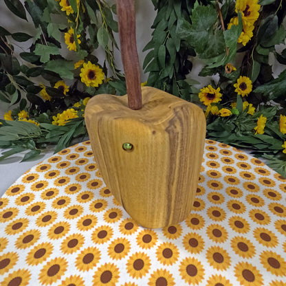 Wooden apple-shaped object on a tablecloth with sunflower pattern, surrounded by greenery and yellow flowers.