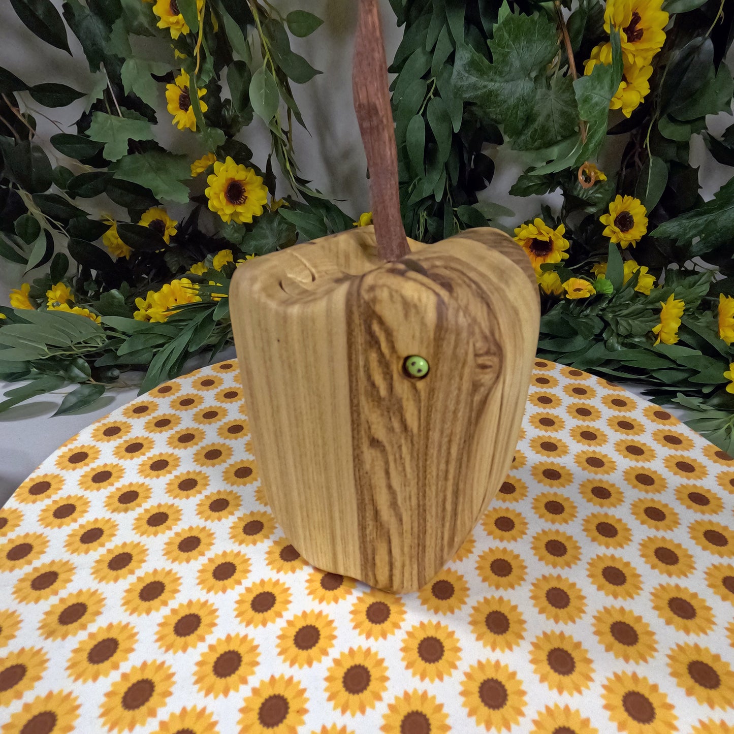 Wooden apple with a handle on a tablecloth with sunflower pattern, surrounded by greenery and yellow flowers.