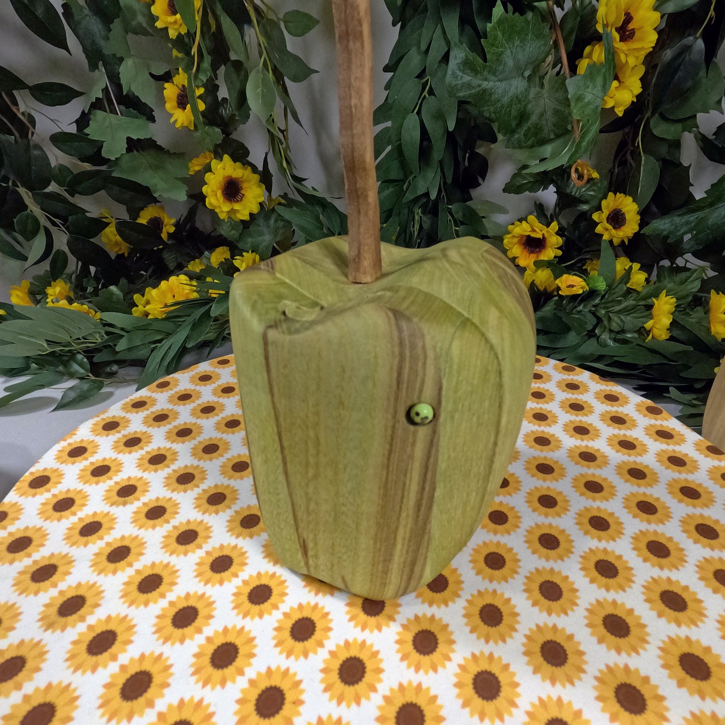 Green wooden apple-shaped object on a tablecloth with sunflower pattern, surrounded by greenery and yellow flowers.