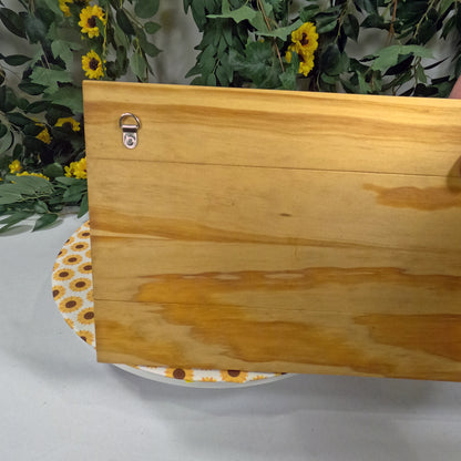 Wooden plaque with floral pattern on a white surface with greenery and yellow flowers in the background