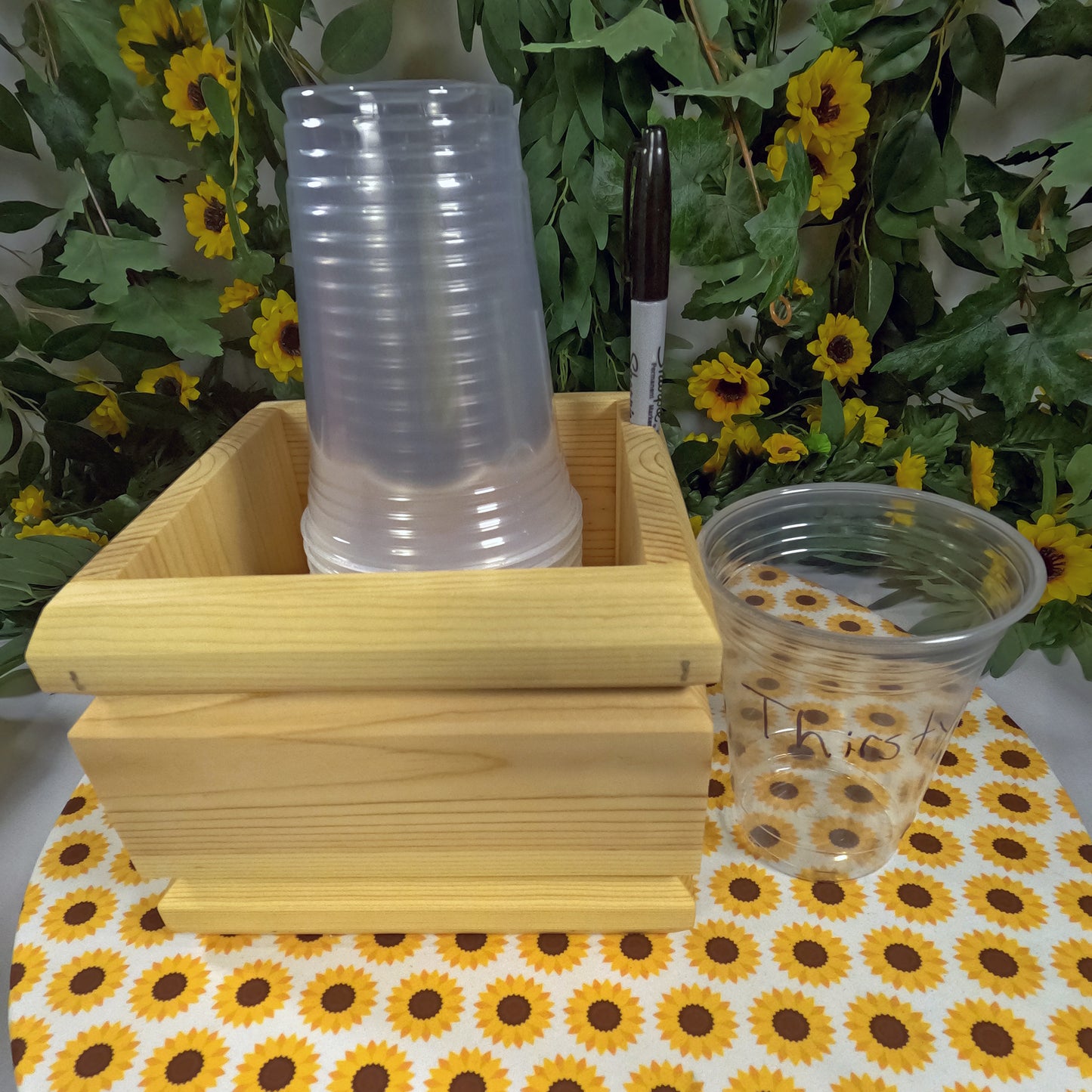 Stack of clear plastic cups in a wooden holder on a tablecloth with sunflower pattern