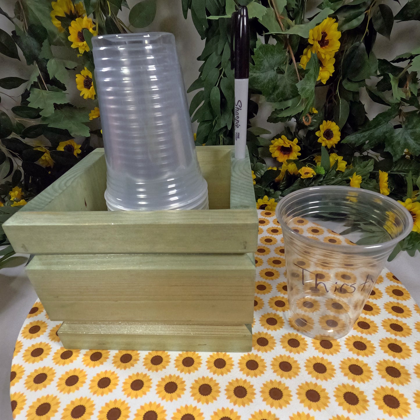 Clear plastic cups on a wooden stand with sunflower-patterned tablecloth and floral background
