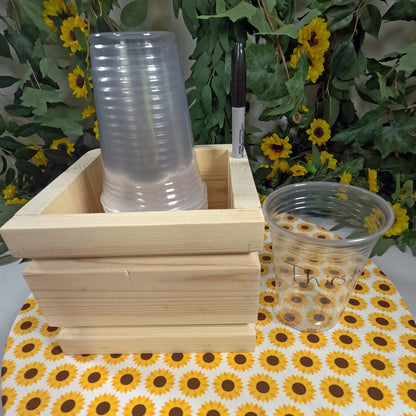 Stack of clear plastic cups in a wooden holder with a pen, on a sunflower-patterned tablecloth.