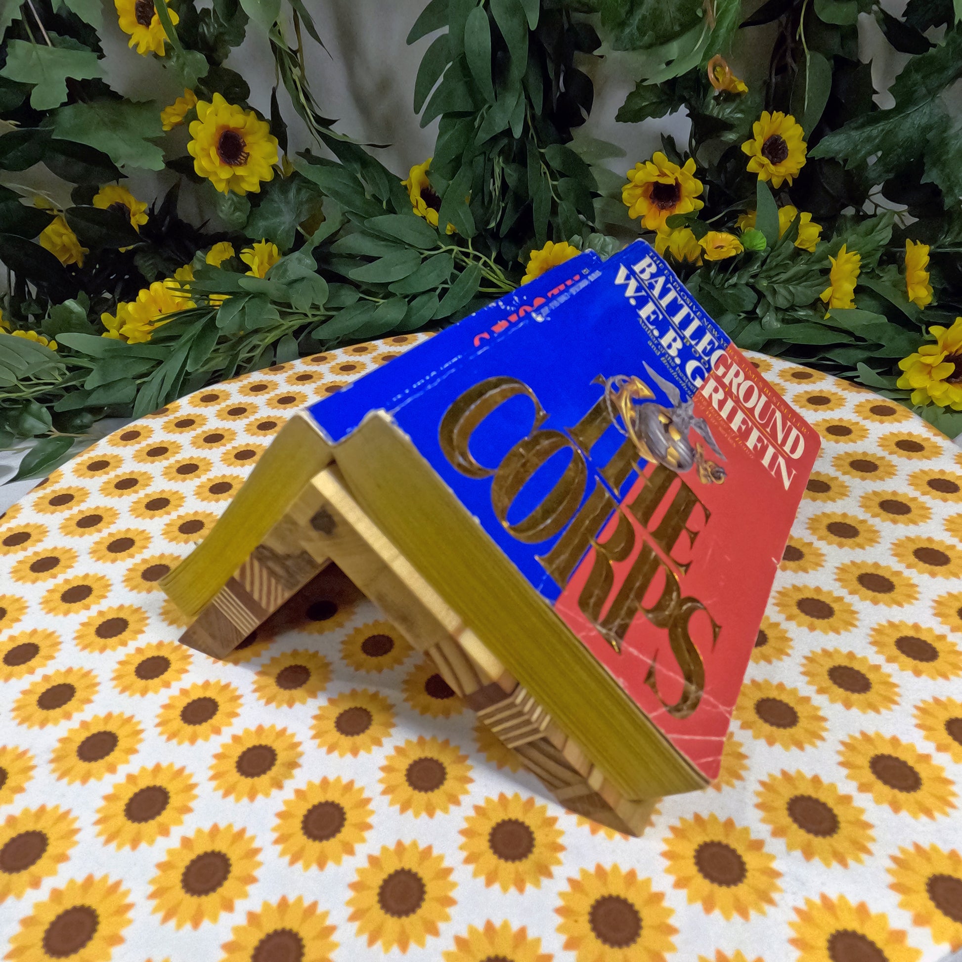 Book titled 'The Corps' on a wooden stand with sunflower-patterned fabric and greenery in the background