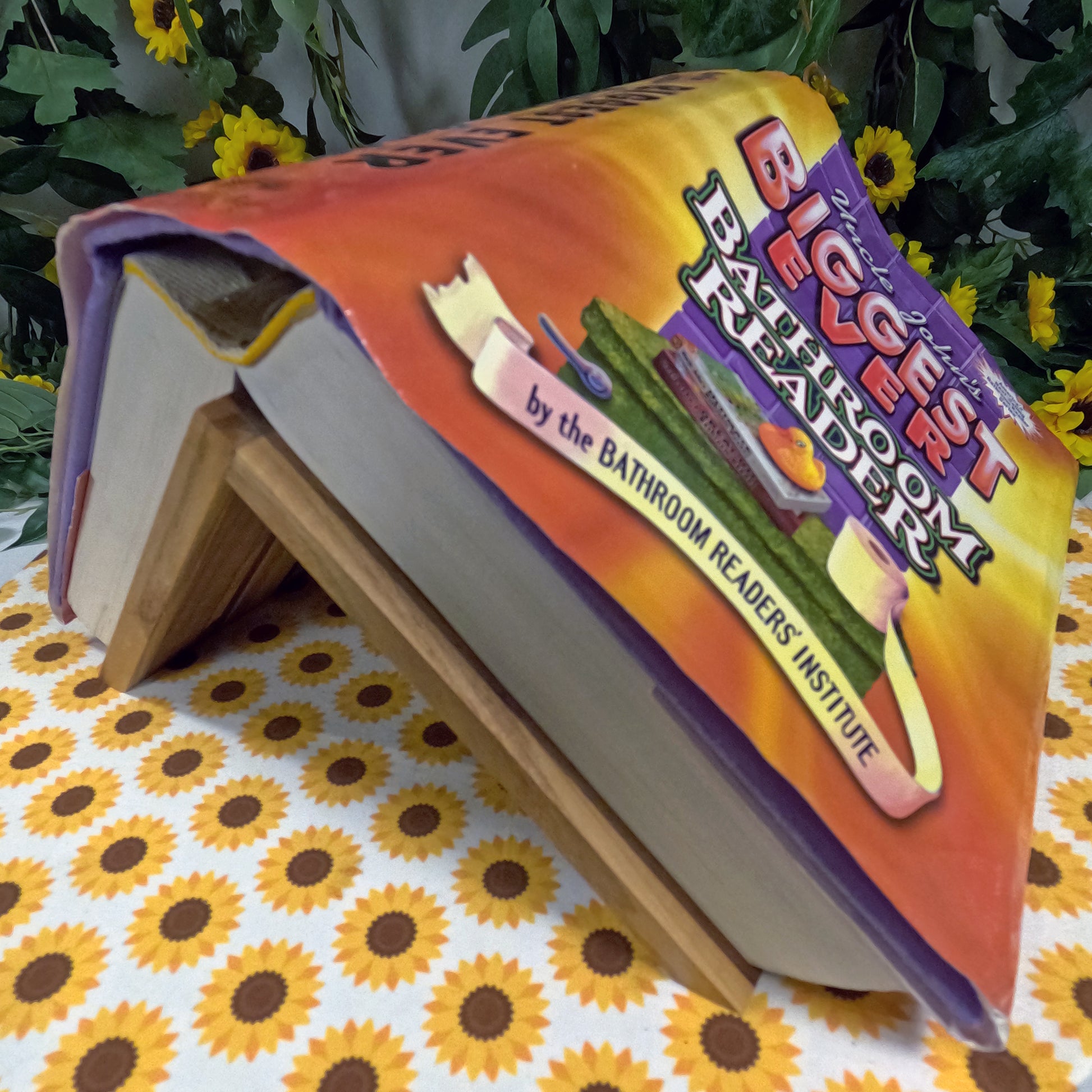 Open book titled 'The Biggest Ever Bathroom Reader' on a floral tablecloth with plants in the background