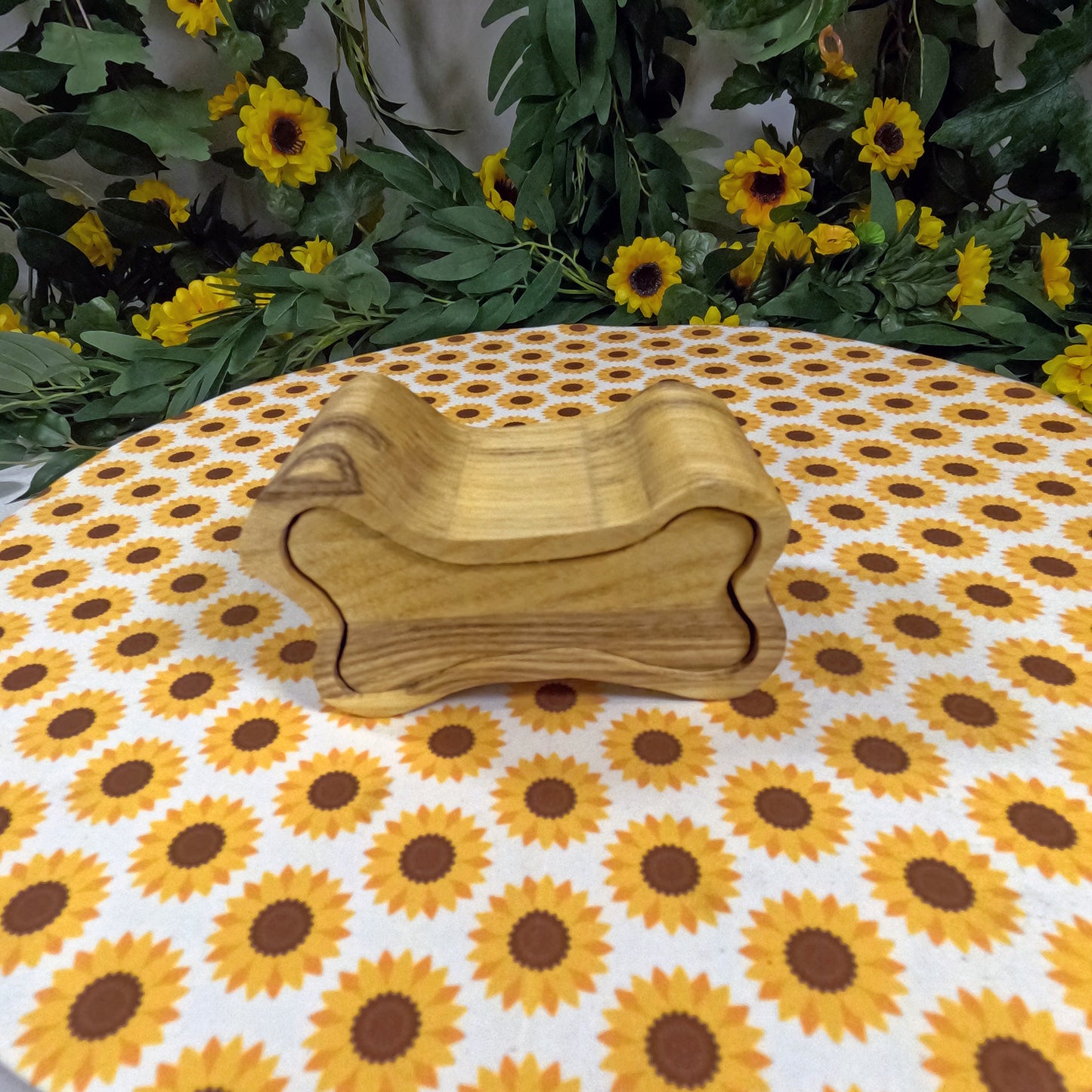 Wooden box on a tablecloth with sunflower pattern, surrounded by greenery and flowers.