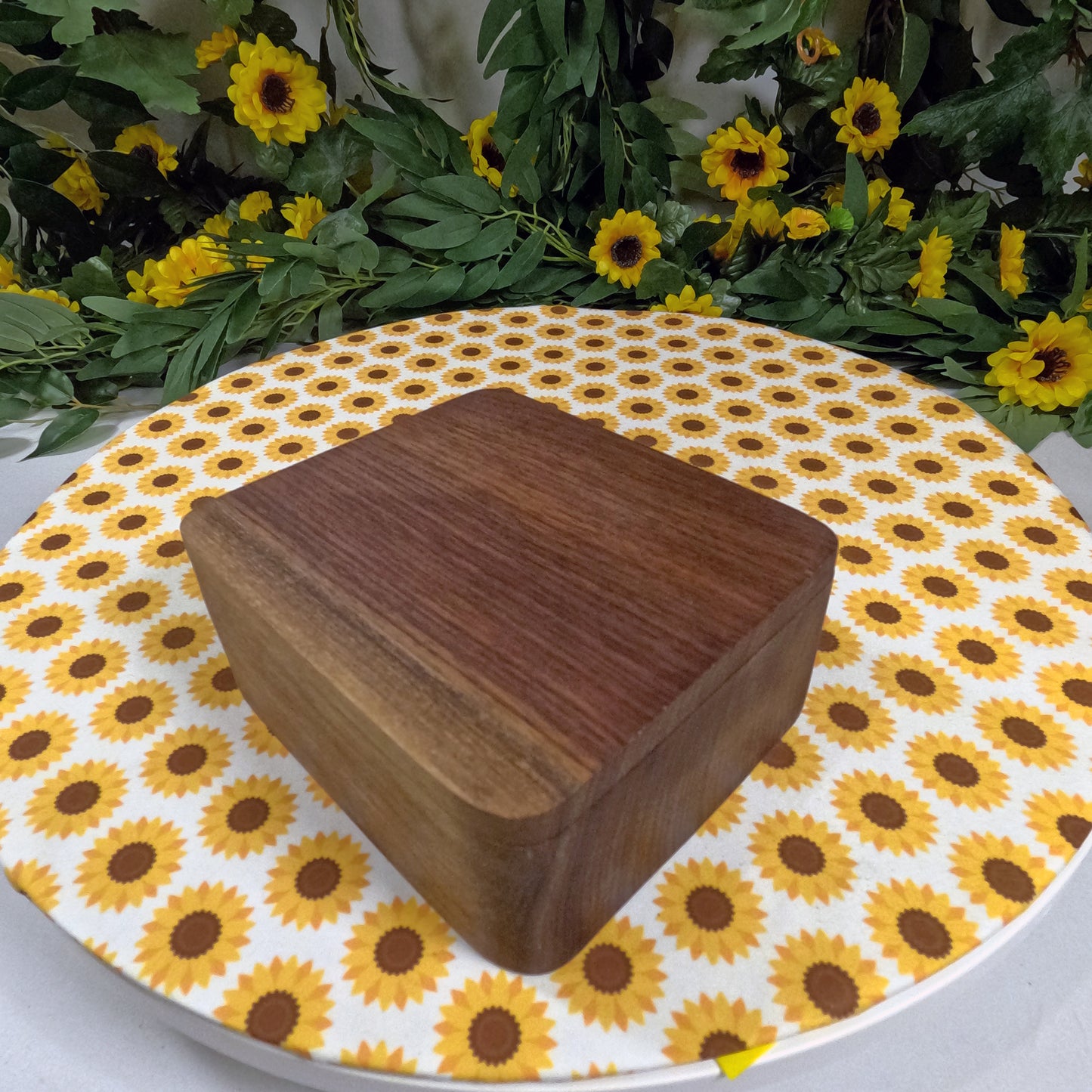 Wooden box on a round table with sunflower pattern, surrounded by greenery and yellow flowers.