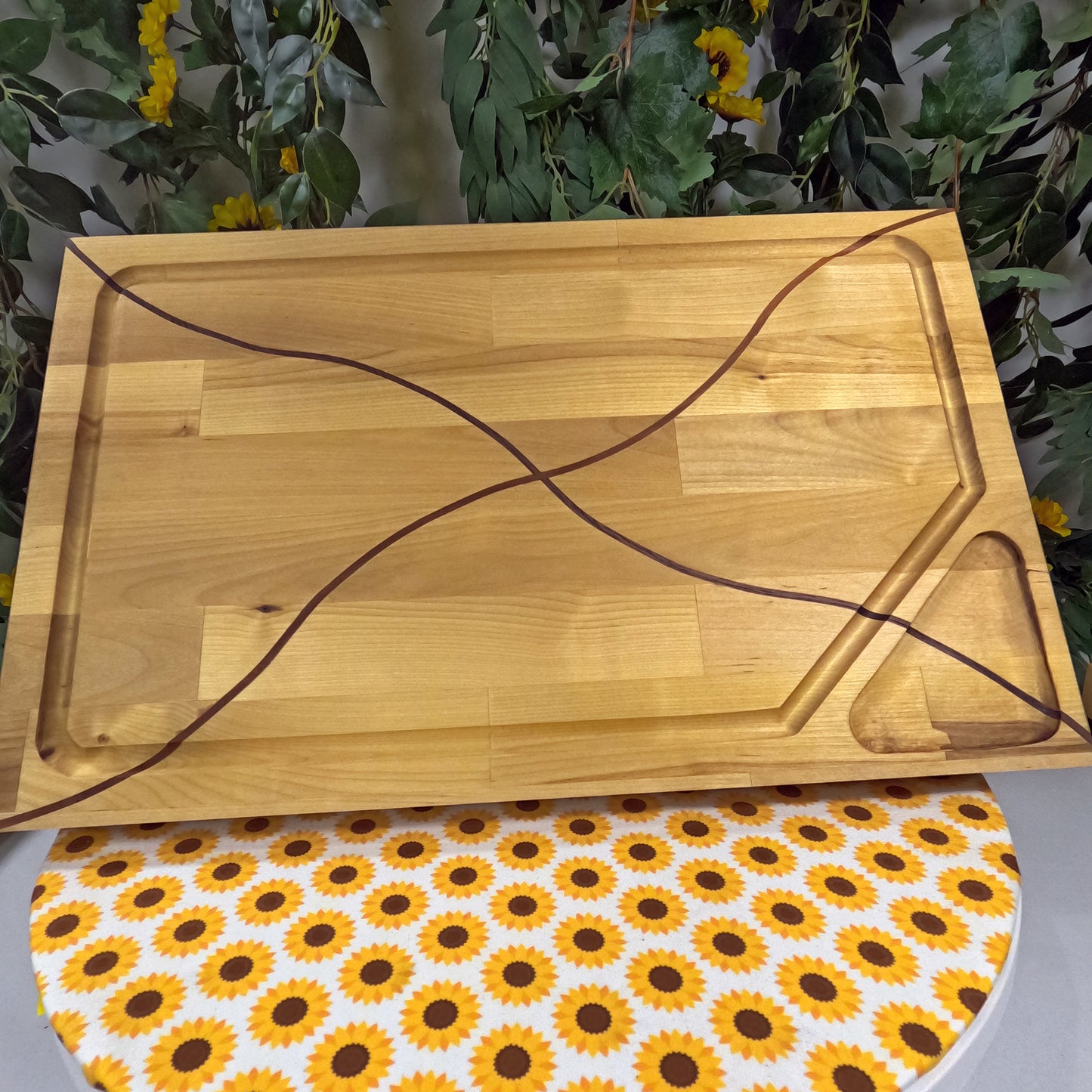 Wooden cutting board on a table with a sunflower patterned mat underneath, surrounded by greenery.