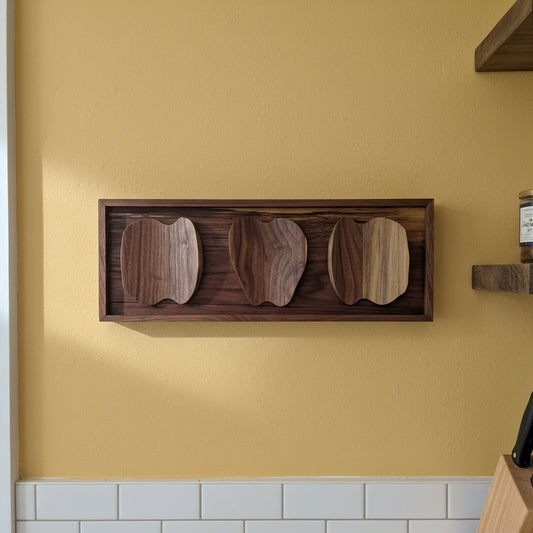 Wooden wall art with three apple cutouts on a white surface with a sunflower-patterned mat underneath, against a green plant background.