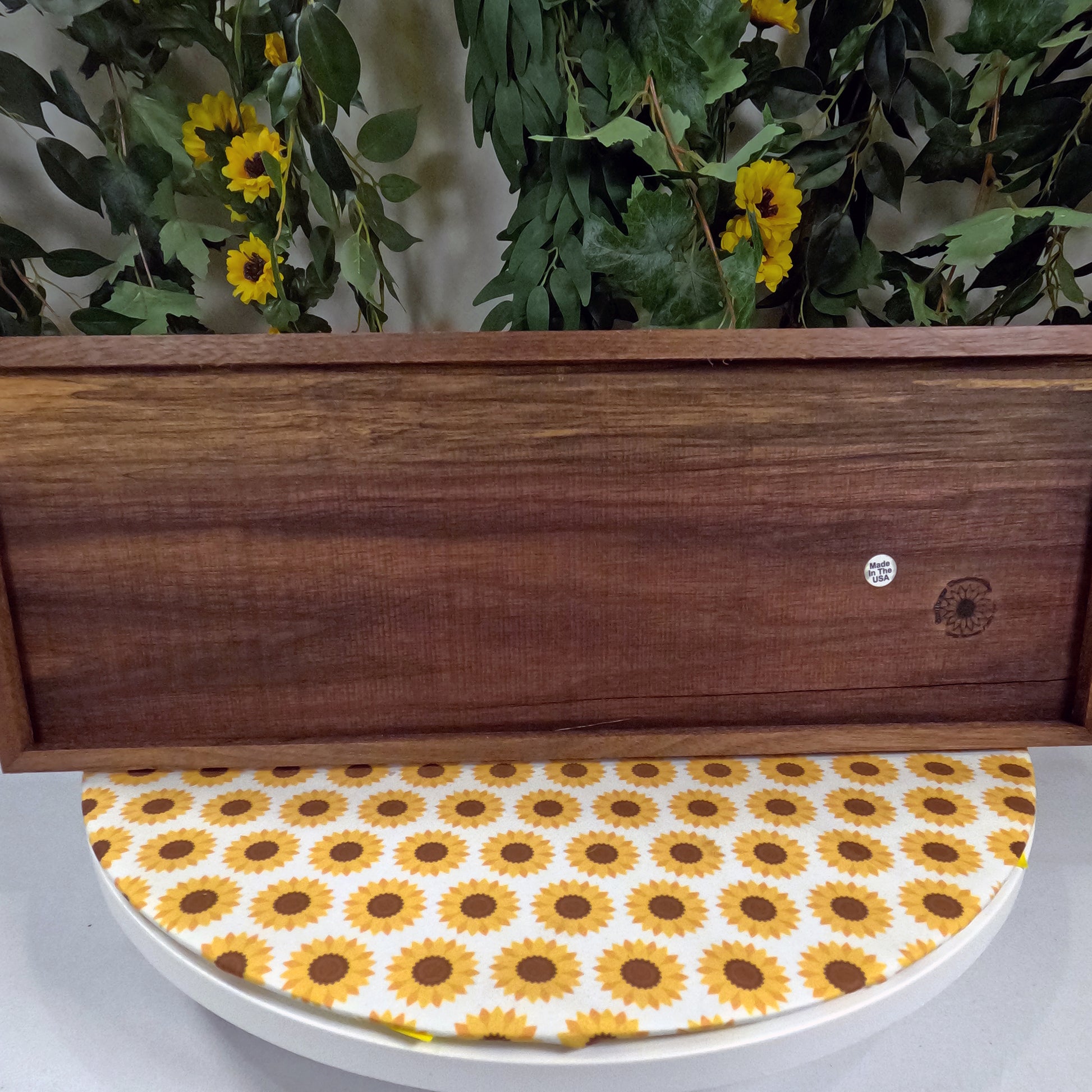 Wooden wall art with sunflower-patterned lid on a white surface, against a background of greenery and yellow flowers.