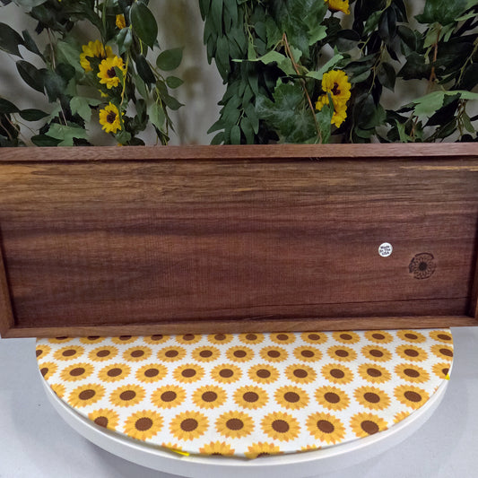 Wooden wall art with sunflower-patterned lid on a white surface, against a background of greenery and yellow flowers.